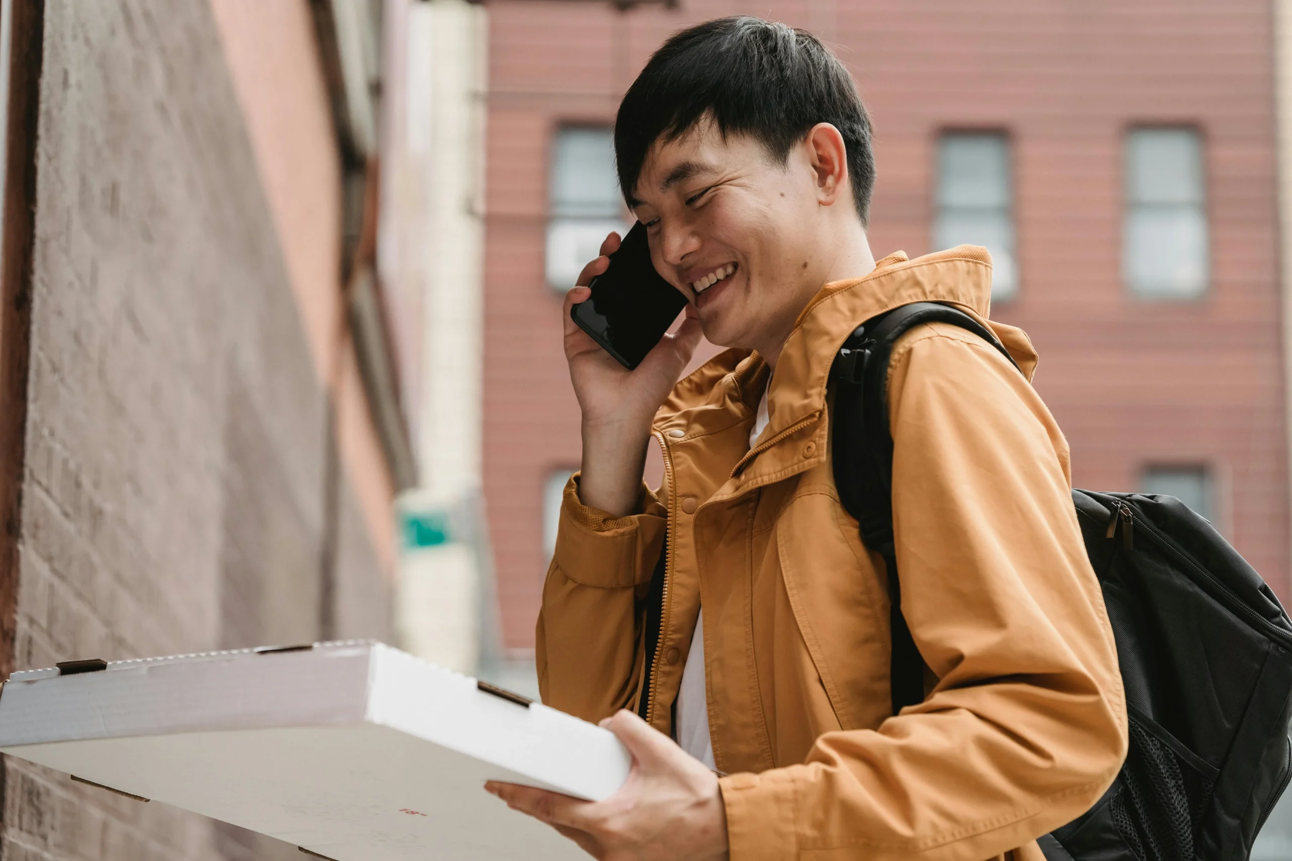 Gen Z financial literacy — Asian male delivery person with pizza box, smiling while on a call in a city street.