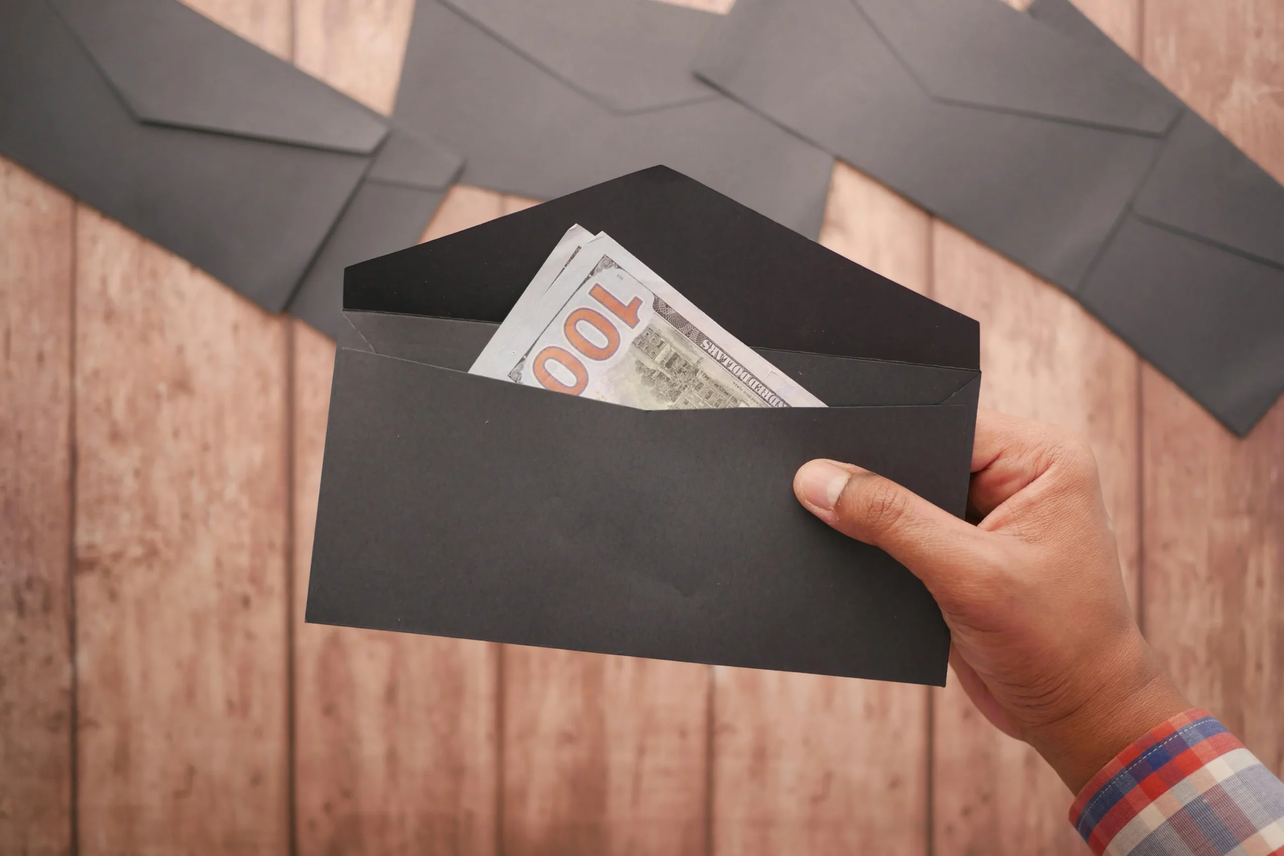 thrifting holiday gifts — A hand holds a black envelope containing a 100 dollar bill on a wooden surface.